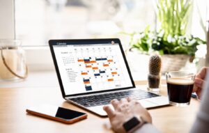 Notebook exibindo um calendário mensal com eventos destacados em laranja e azul-escuro sobre uma mesa de trabalho, ao lado de um café, um smartphone e plantas verdes ao fundo.